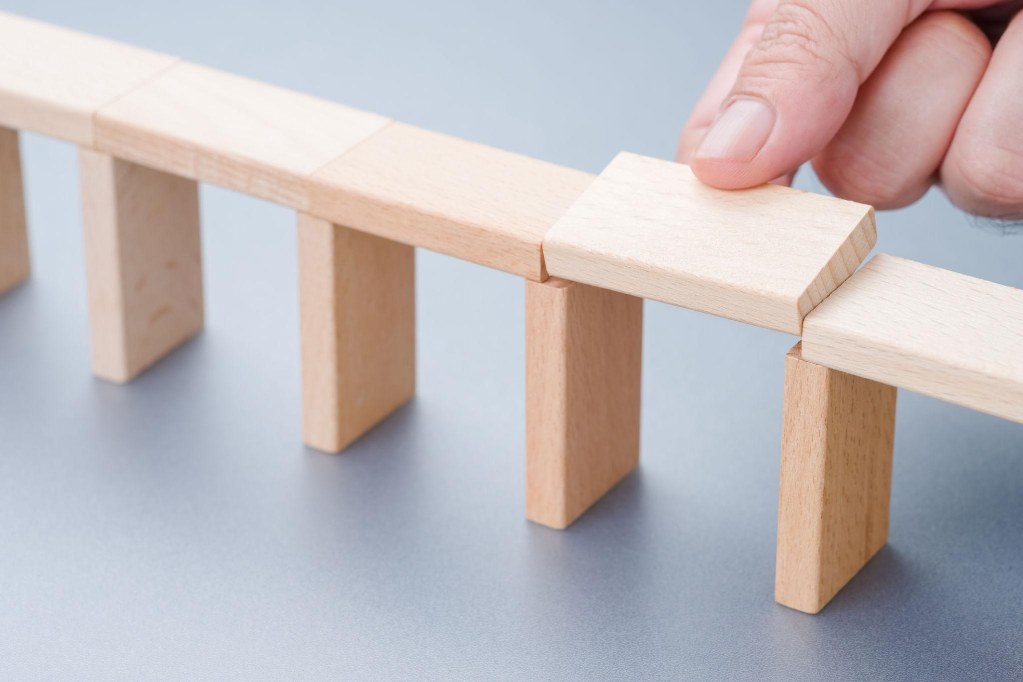 Persoon voegt een houten bouwsteen toe aan een constructie, een metafoor voor het opbouwen van spreekvaardigheid en zelfvertrouwen in kleine stappen.